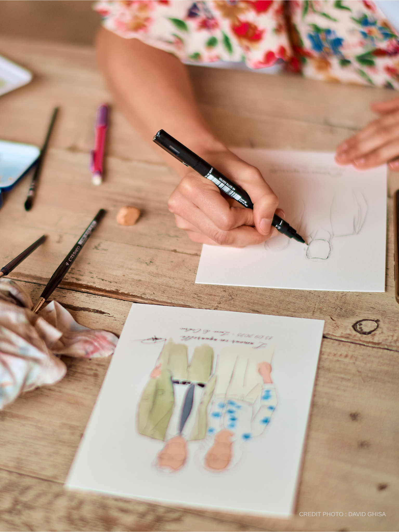 Une personne dessine ou peint un modèle de vêtement, probablement une robe, sur une feuille de papier avec des crayons ou des feutres. Des accessoires d'art sont dispersés autour de la feuille, sur une surface en bois.