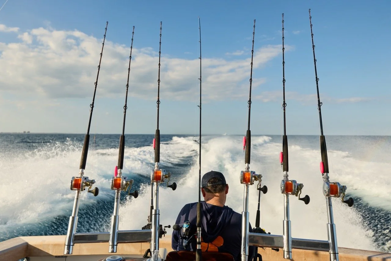 A person wearing a dark hoodie and cap sitting on a boat, fishing with six fishing rods mounted at the back of the boat, with a view of the open sea and a partly cloudy sky in the background.