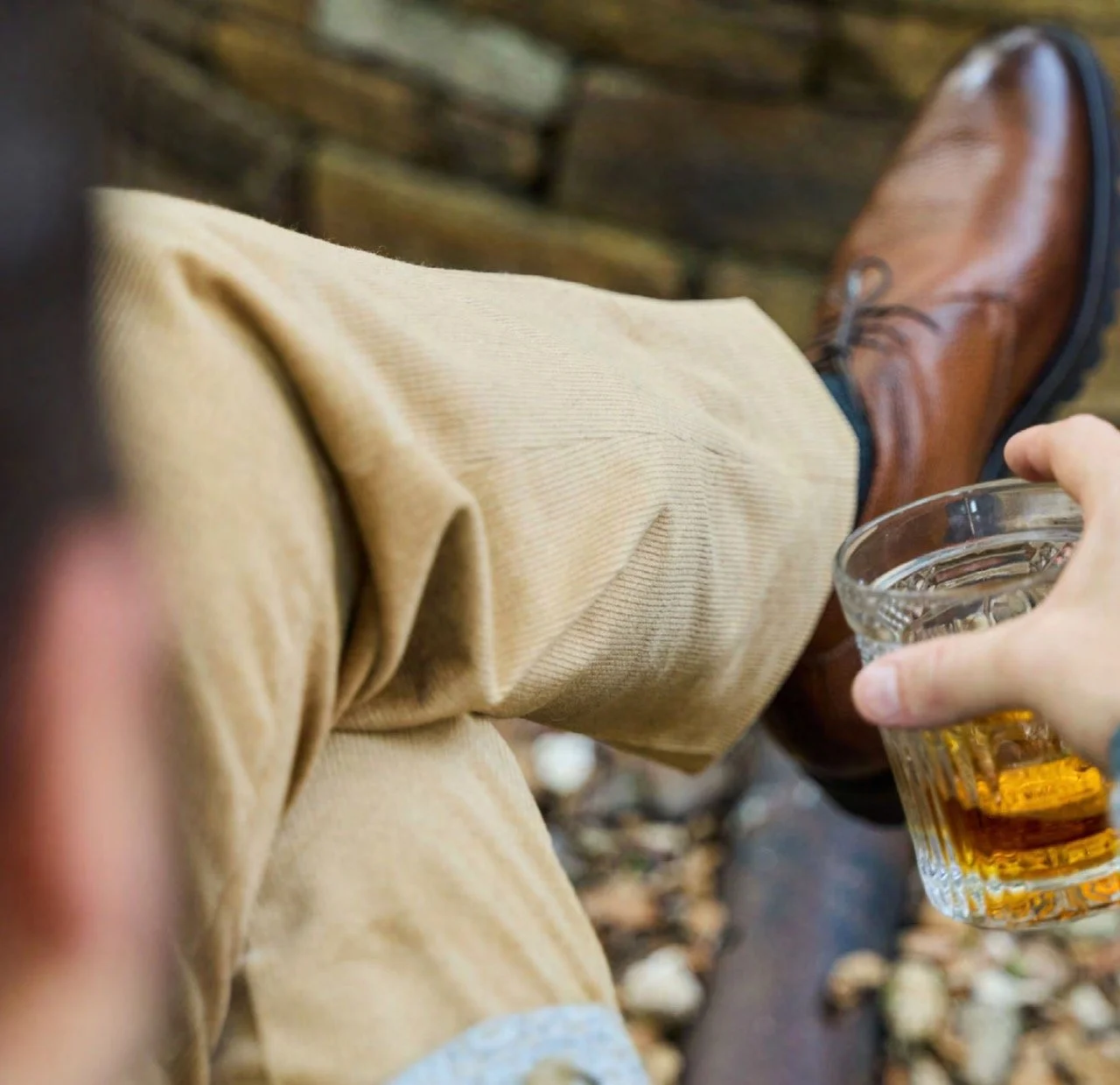 Person sitting on a wooden bench, wearing tan pants and brown leather boots, holding a glass of beer.