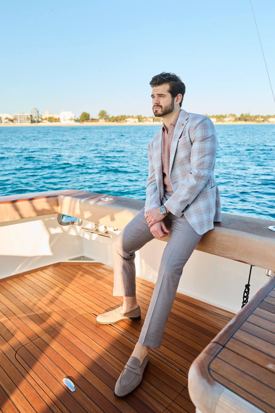 Man wearing a mauve sharkskin Malcom Como trousers with a mauve short-sleeve knit shirt and a bold plaid sportscoat seated at the back of a yacht looking out over the water. 