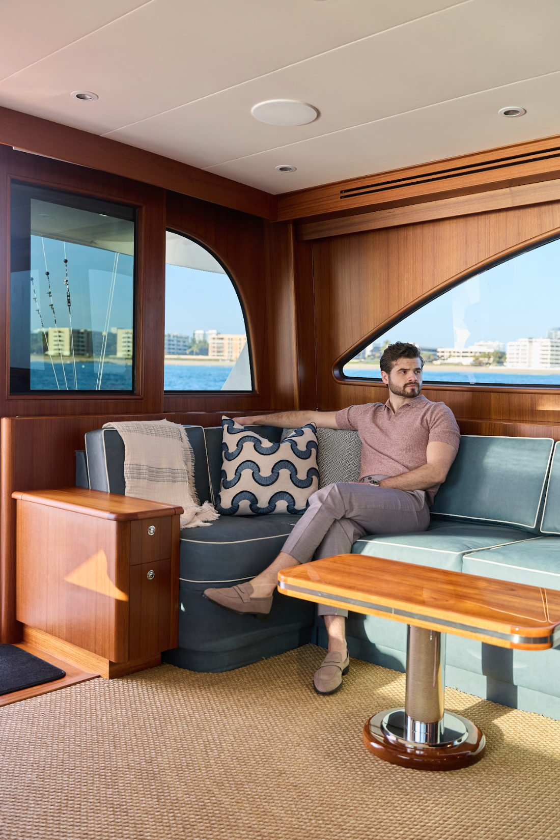  Man dressed in Malcom’s Como model trouser with a mauve short-sleeve polo and loafers seated in the interior salon of a yacht looking out the window. 