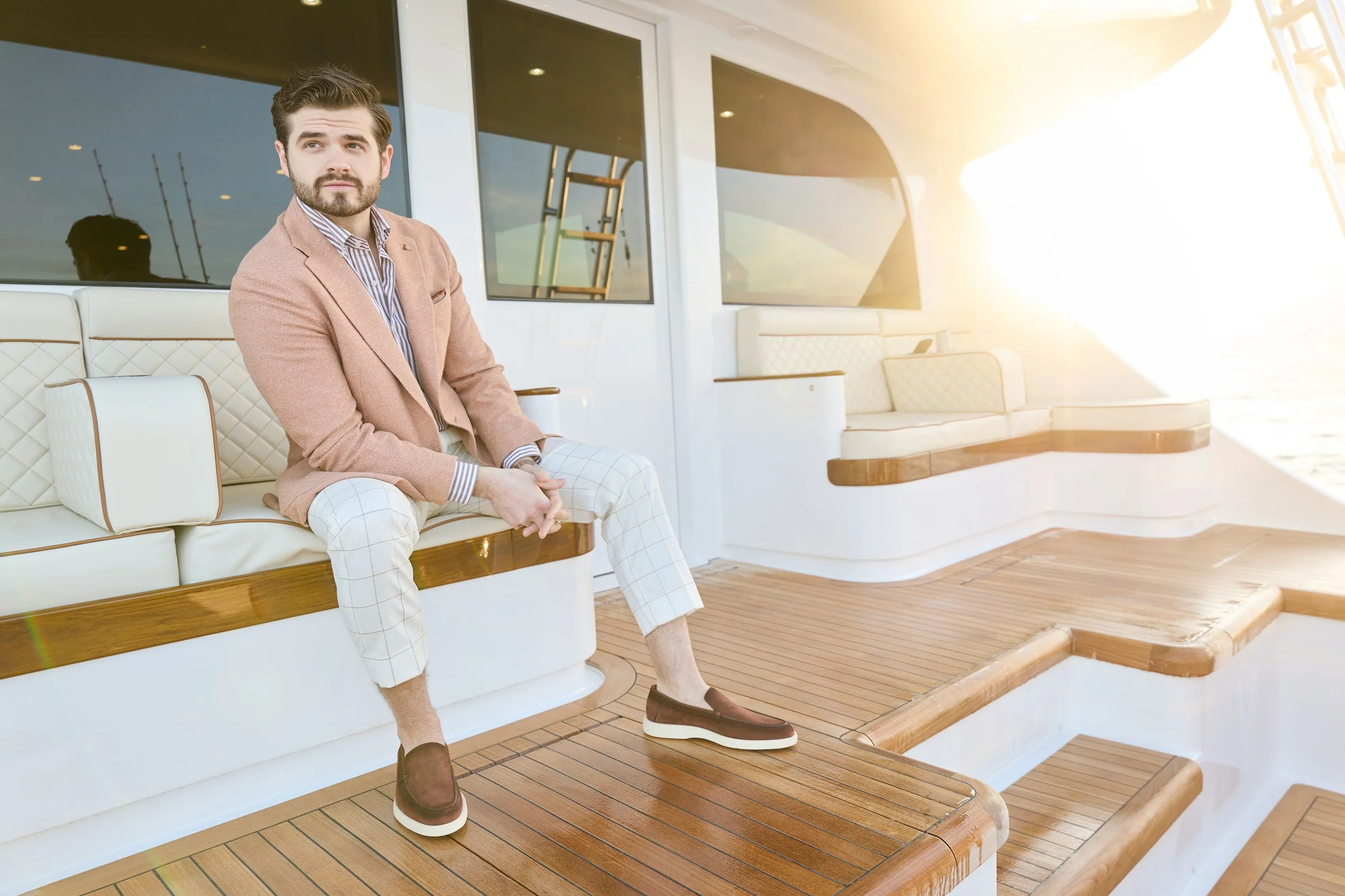  Man wearing Malcom’s Padova model trouser in cream with a brown windowpane pattern with a brown bengal stripe dress shirt and a soft mauve blazer with loafers seated at the back of a yacht watching the sunset. 