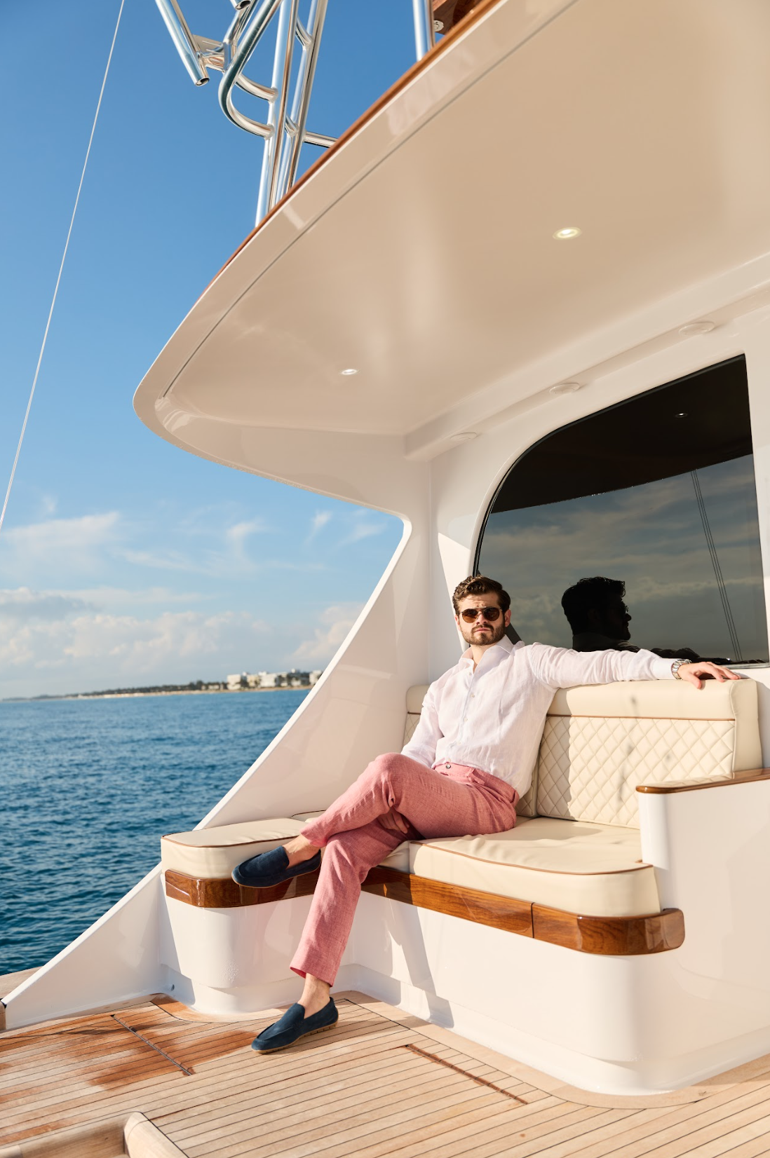  A man wearing a pink linen Malcom Besano model 5-pocket luxury trousers with a long-sleeve white linen shirt and blue loafers sitting in the sun at the back of the yacht.  