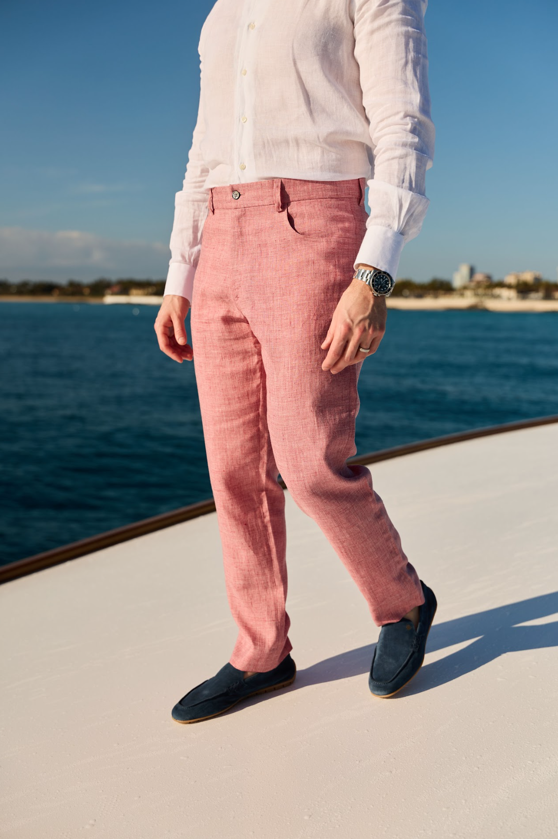  Man wearing pink linen Malcom Besano model 5-pocket trousers with a white linen long-sleeve shirt and blue loafers walking along the front of the yacht.  
