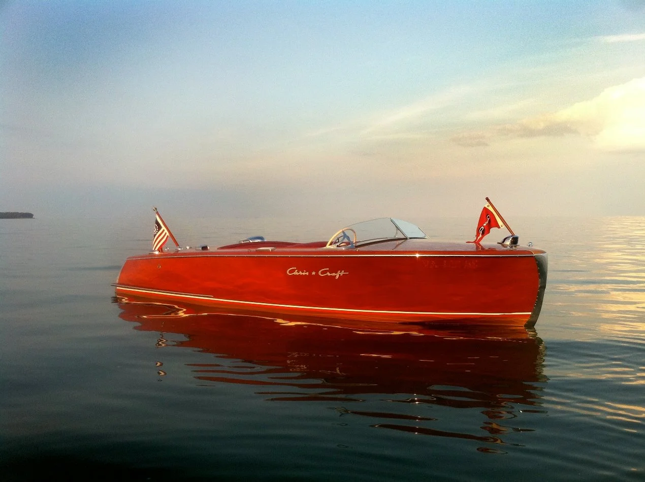 A vintage wooden Chris Craft speedboat floating on calm water at sunset with a vintage American flag off the stern and a nautical flag at the bow waving against a clear sky in the background.