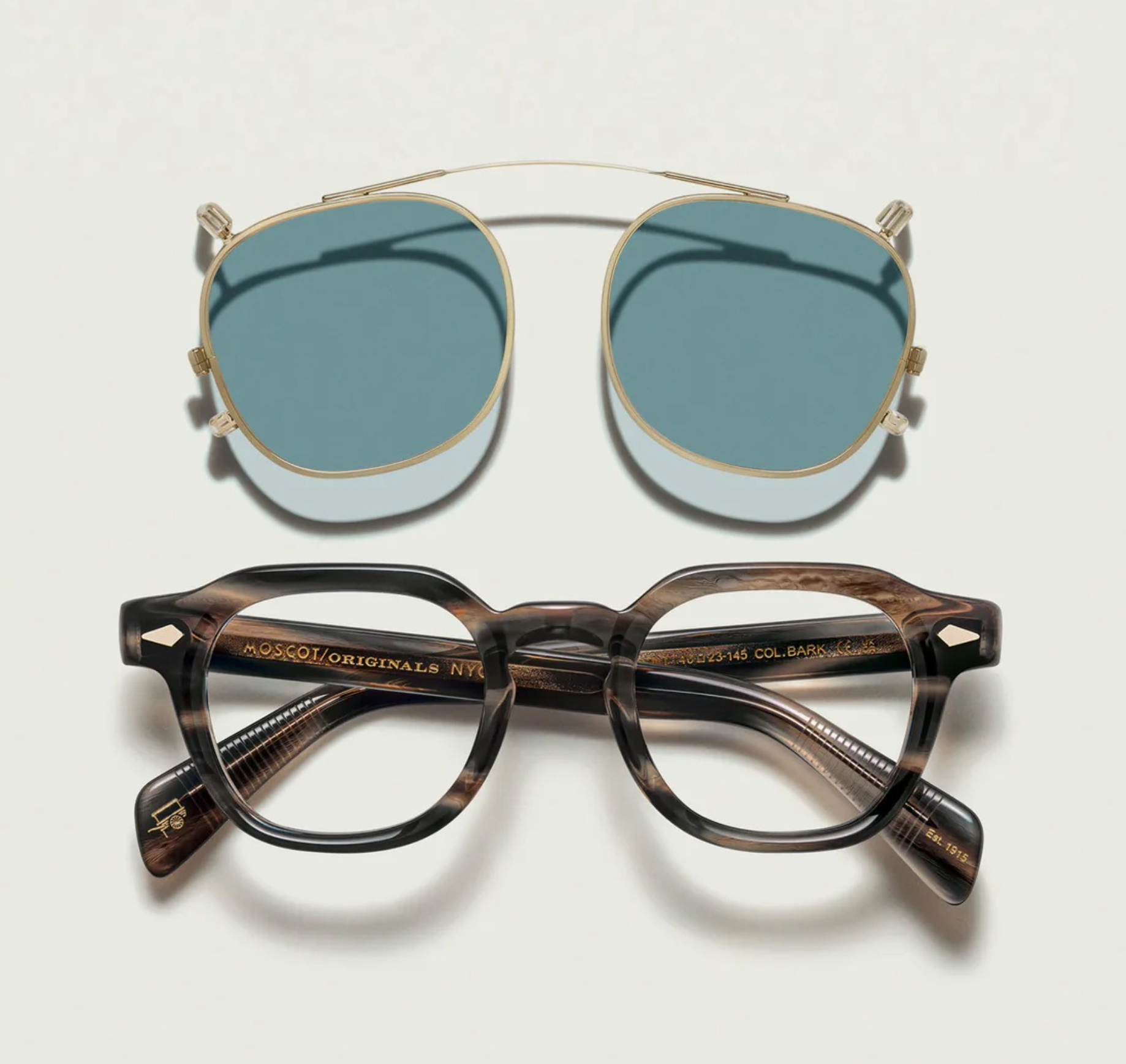 A pair of Moscot sunglasses with reflective blue lenses, a pair of Moscot eyeglasses with tortoiseshell frames placed on a white surface casting deep shadows.