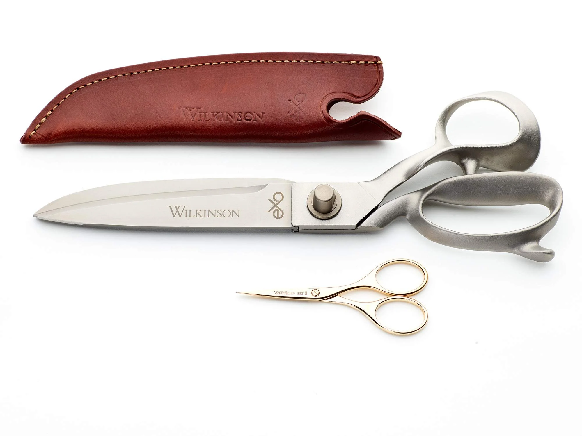 A pair of industrial silver scissors with a curved handle, a gold ring, and a leather case with the brand name 'Wilkinson' embossed on it place above a small pair of Whiteley trimming scissors.
