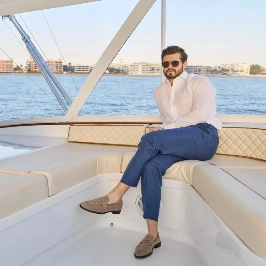 Man with sunglasses and beard sitting on boat with water and city in background.