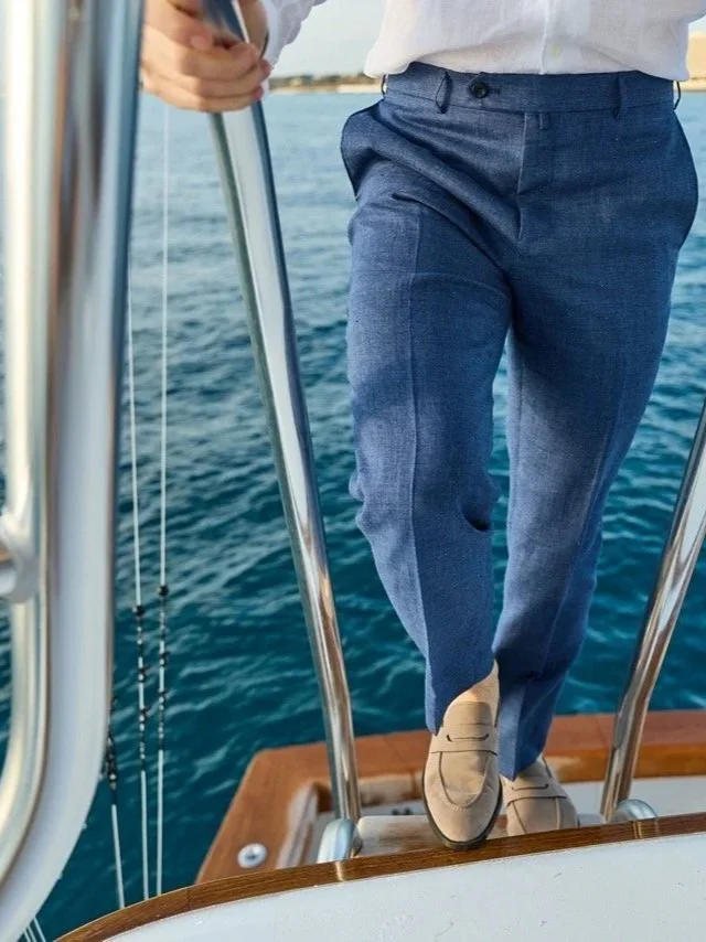 A person standing on the deck of a boat, wearing beige shoes, blue dress pants, and a white shirt, with water and shoreline in the background.