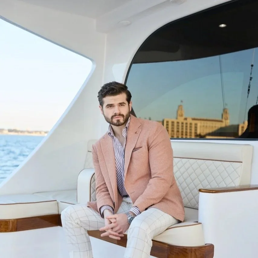 A man with dark hair and a beard, wearing a peach blazer and white checkered pants, sitting on a yacht with water and a cityscape in the background.