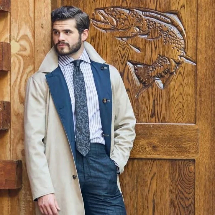 A man with dark hair and a beard, dressed in a blue striped shirt, patterned tie, dark pants, and a beige coat, standing against a wooden background with carved animals.