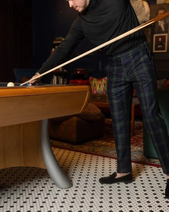 A person in tartan pants and black top is playing pool, leaning over and holding a cue stick aimed at the cue ball on a wooden pool table.