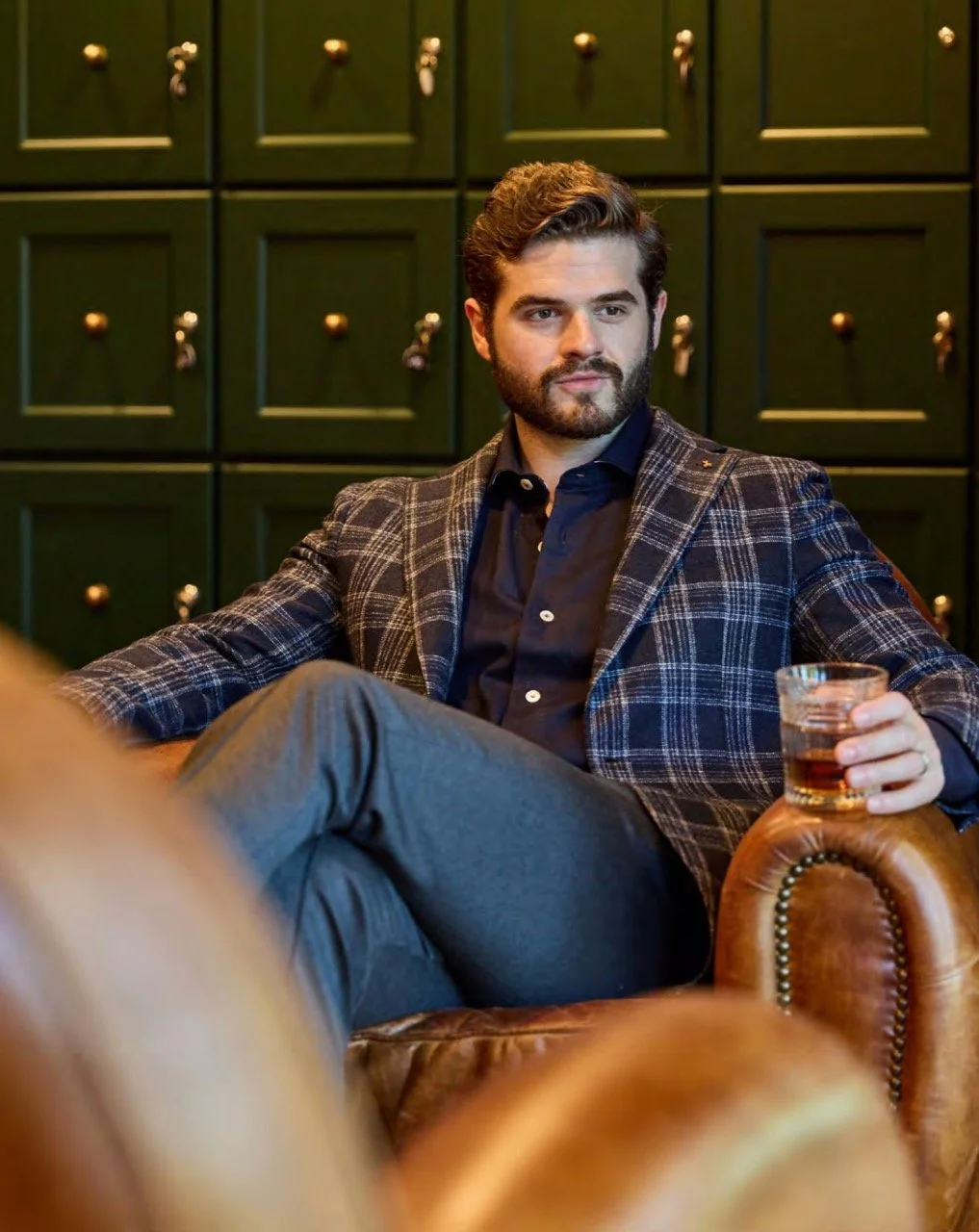 A man with a beard and wavy hair sitting on a brown leather chair, holding a glass of dark beverage, in front of a green wall with small drawers.