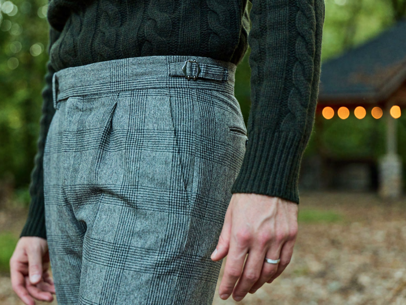 Close-up of a person wearing plaid gray trousers and a dark green cable-knit sweater, standing outdoors with greenery and string lights in the background.