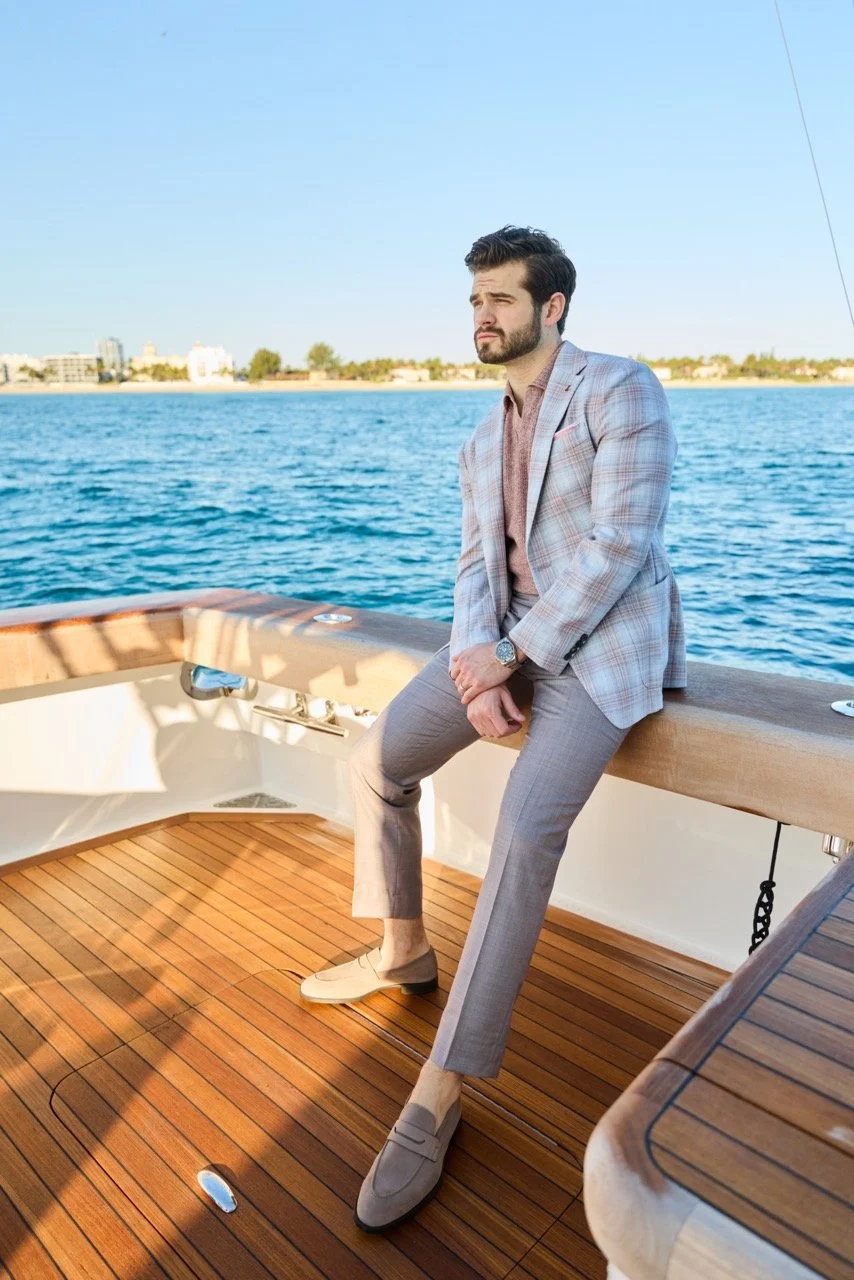 A man in a plaid blazer and gray trousers sitting on the edge of a boat, overlooking the water with a city skyline in the background.