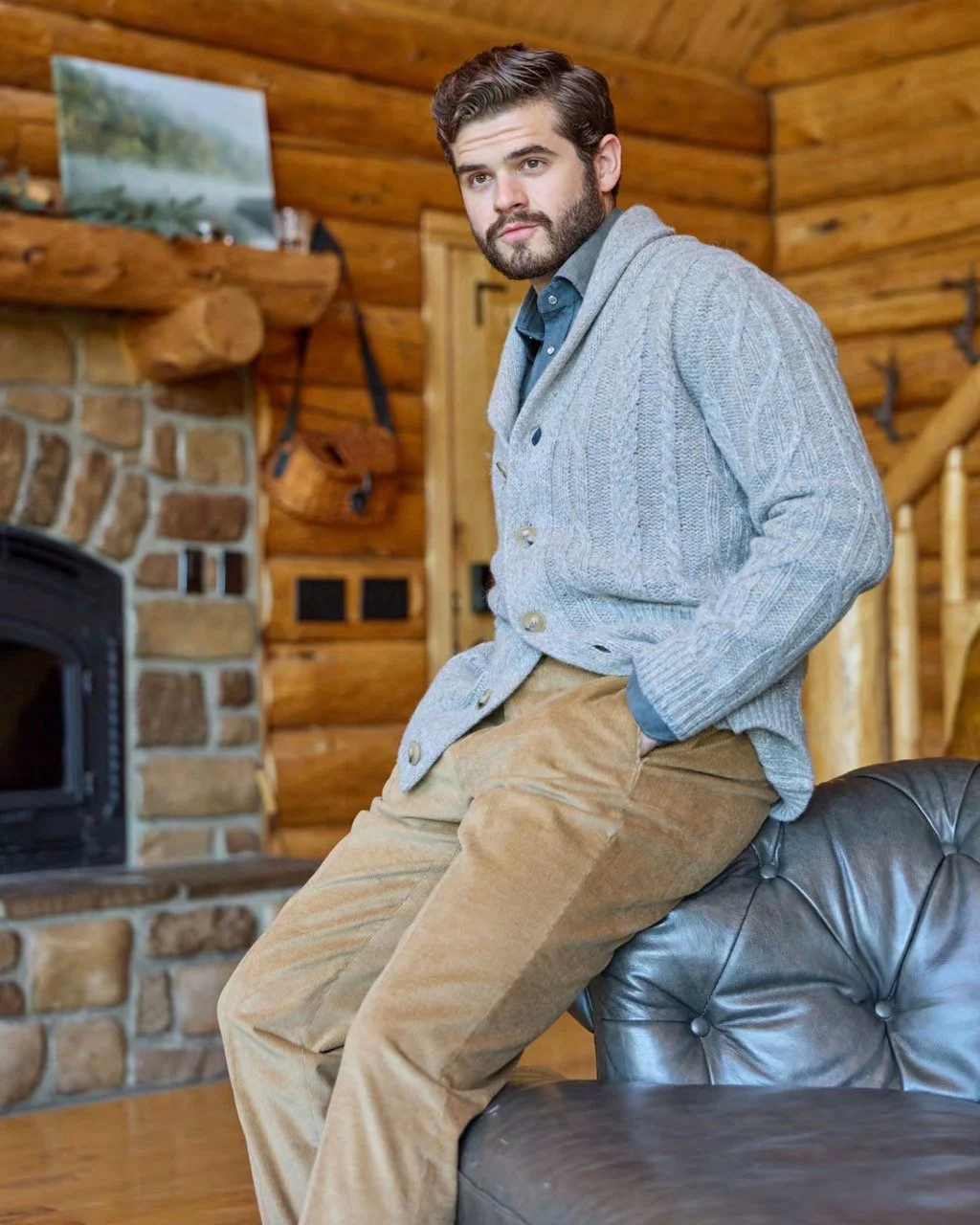 A man wearing a blue shirt, gray sweater, and tan corduroy pants, sitting on the arm of a black leather sofa in a cozy, log cabin-style room with wooden walls and a stone fireplace.