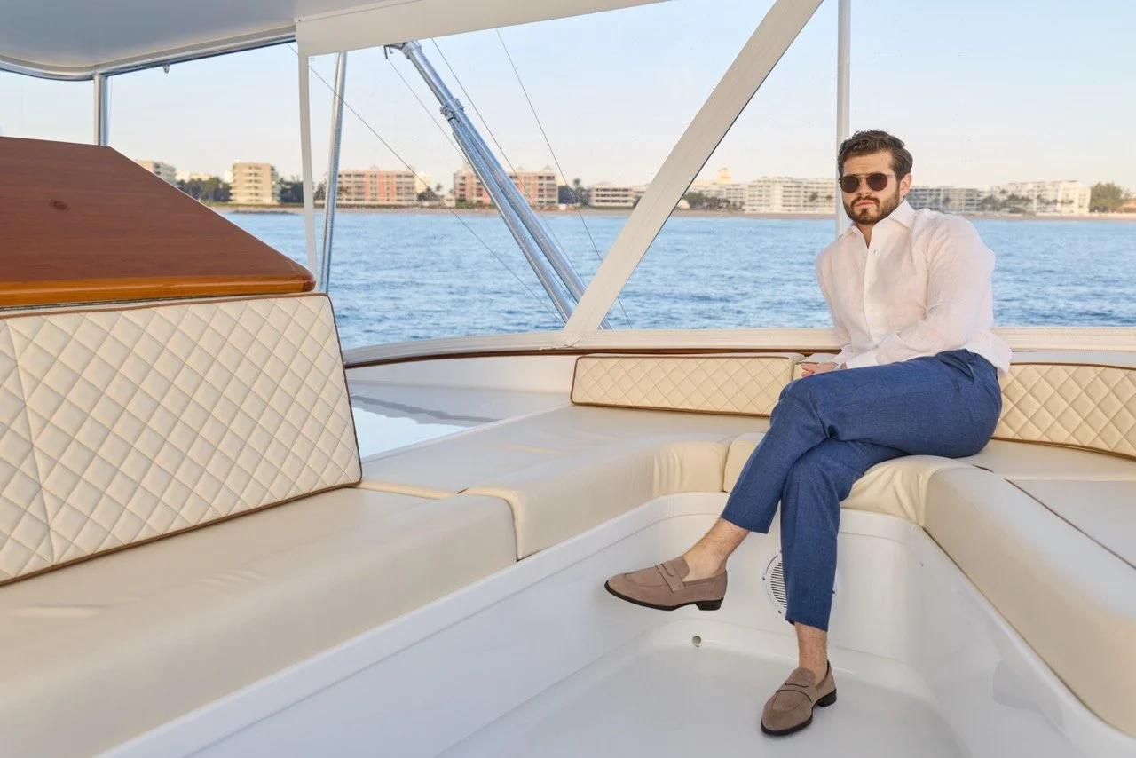 Man sitting on a white cushioned seat on a yacht, wearing sunglasses, a white shirt, blue linen Malcom Parma trousers, and brown loafers, with water and buildings in the background.