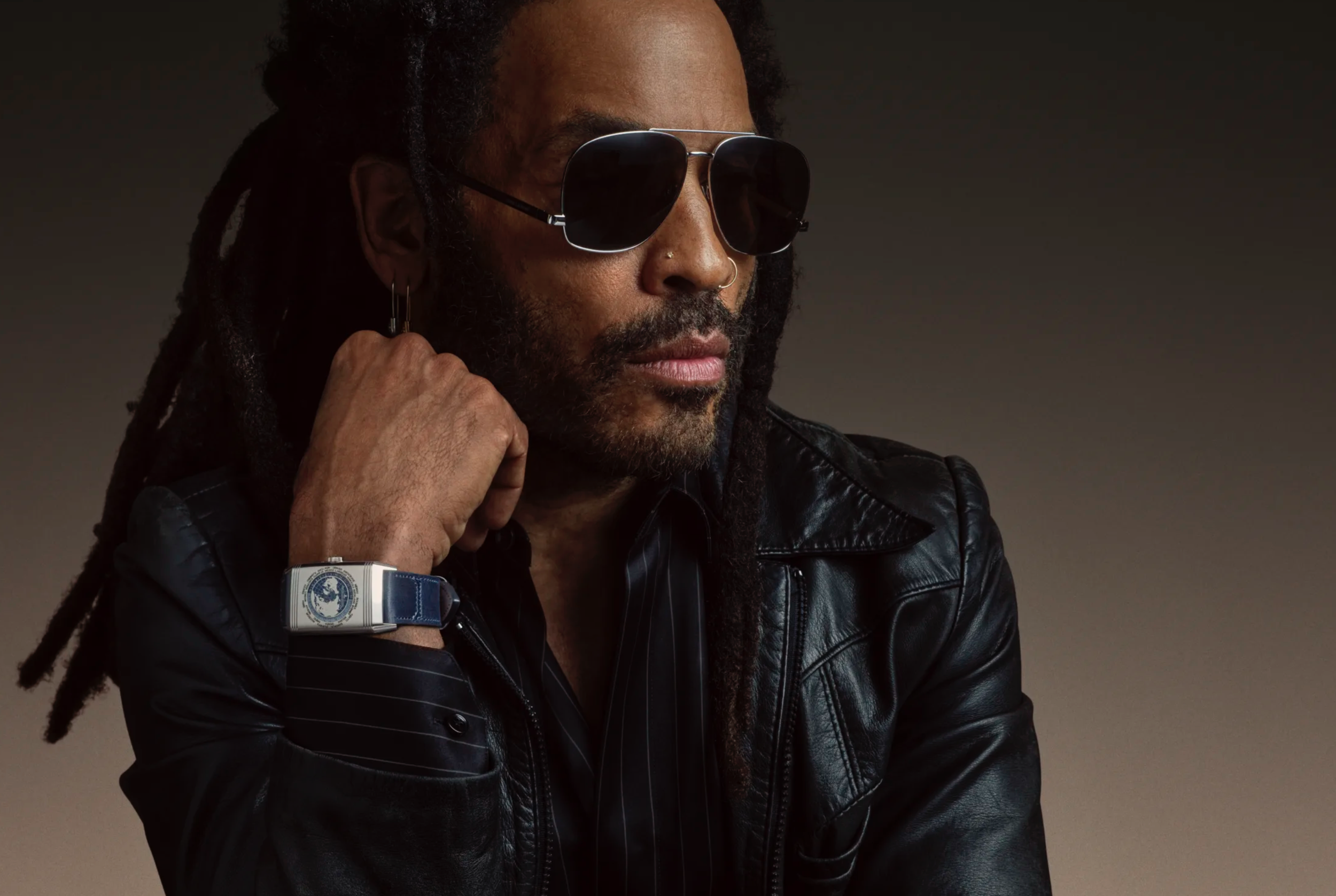Lenny Kravitz wearing a black leather jacket over a black pinstriped shirt and dark aviator sunglasses sporting a JaegerLeCoultre Geographique watch in silver and blue.