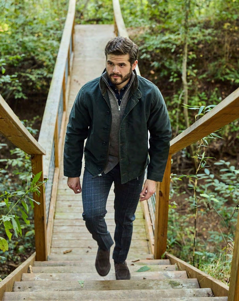 A man walking up wooden stairs on a forested pathway, wearing a dark jacket, plaid pants, and brown shoes.