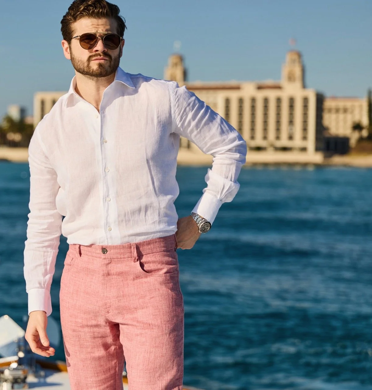 A man with dark hair and a beard wearing sunglasses, a white linen shirt, pink pants, and a silver watch standing by water with a cityscape in the background.
