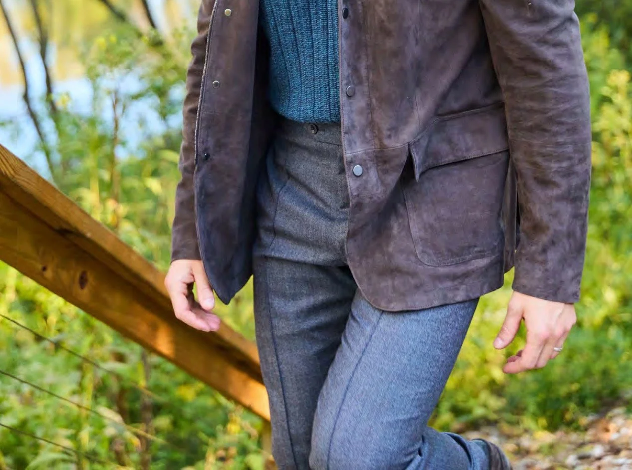 Close-up of a person wearing a brown leather jacket, blue sweater, and gray pants, walking on a wooded trail with a wooden railing nearby.