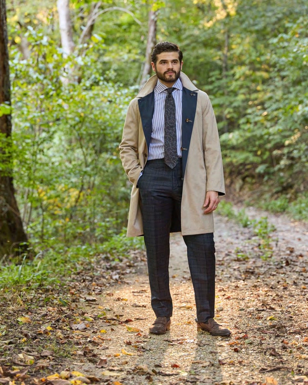 A man dressed in a business suit and overcoat standing on a forest trail surrounded by green trees.