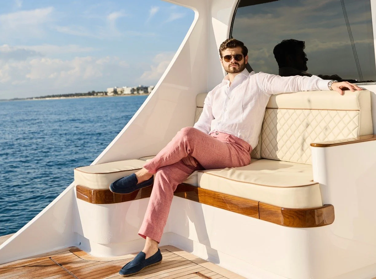 A man wearing sunglasses, a white shirt, pink pants, and navy loafers is sitting on a cream leather and wood bench on a yacht, with water and a distant shoreline in the background.