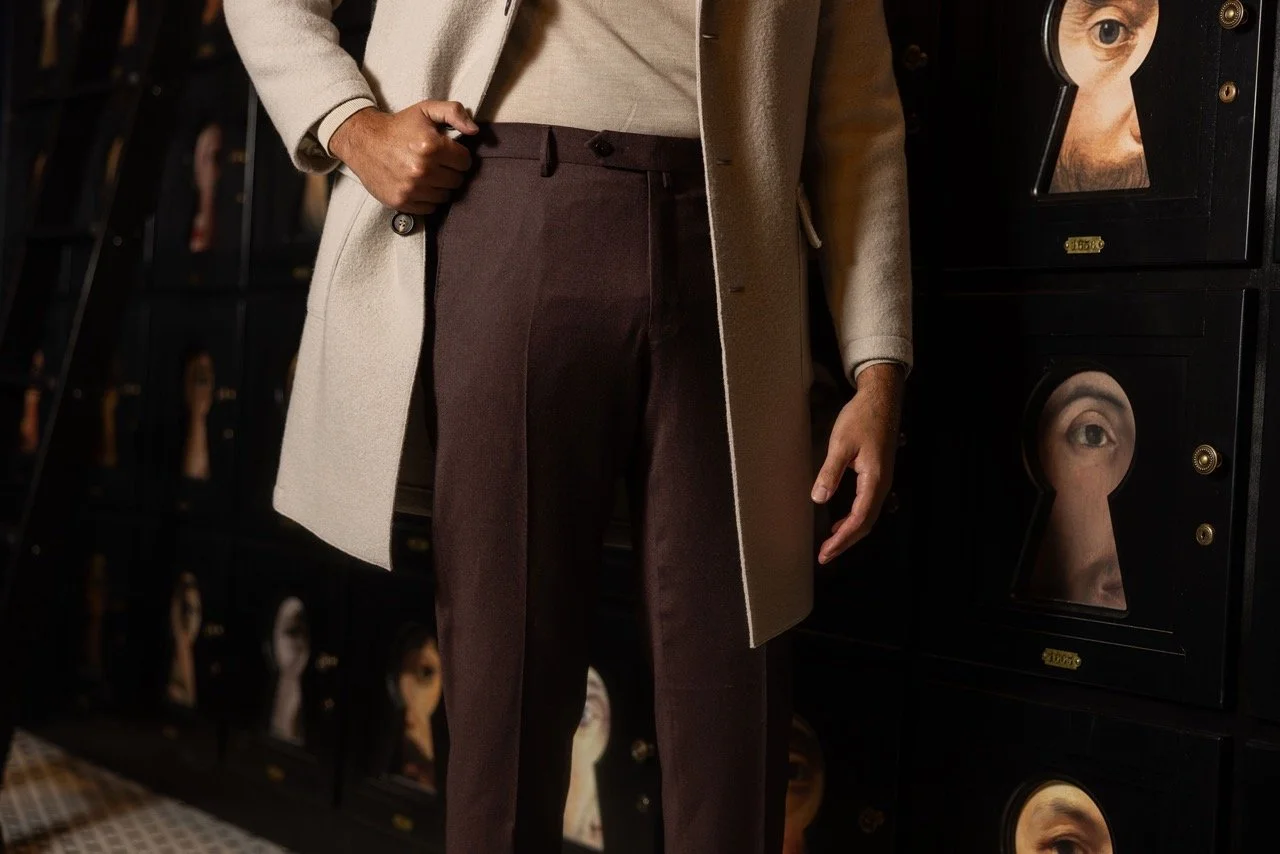 Person wearing a beige coat and dark brown pants, standing next to a wall of black lockers with eyes behind keyhole windows.