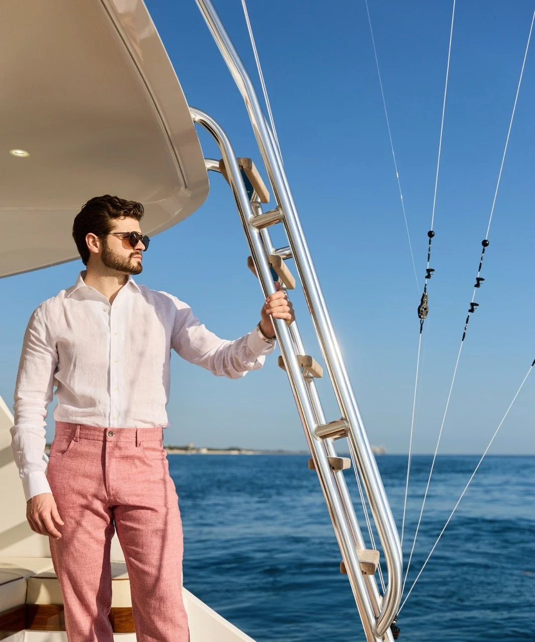 Man wearing white shirt and pink pants holding a boat railing on a yacht with a blue ocean and sky in the background.