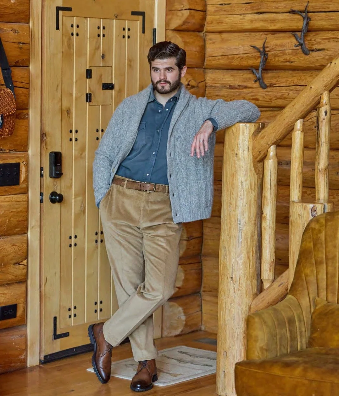A man with a beard and dark hair, dressed in a blue shirt, beige pants, and brown boots, stands inside a log cabin next to a wooden staircase. He leans on the staircase railing with his left arm and looks to the side.