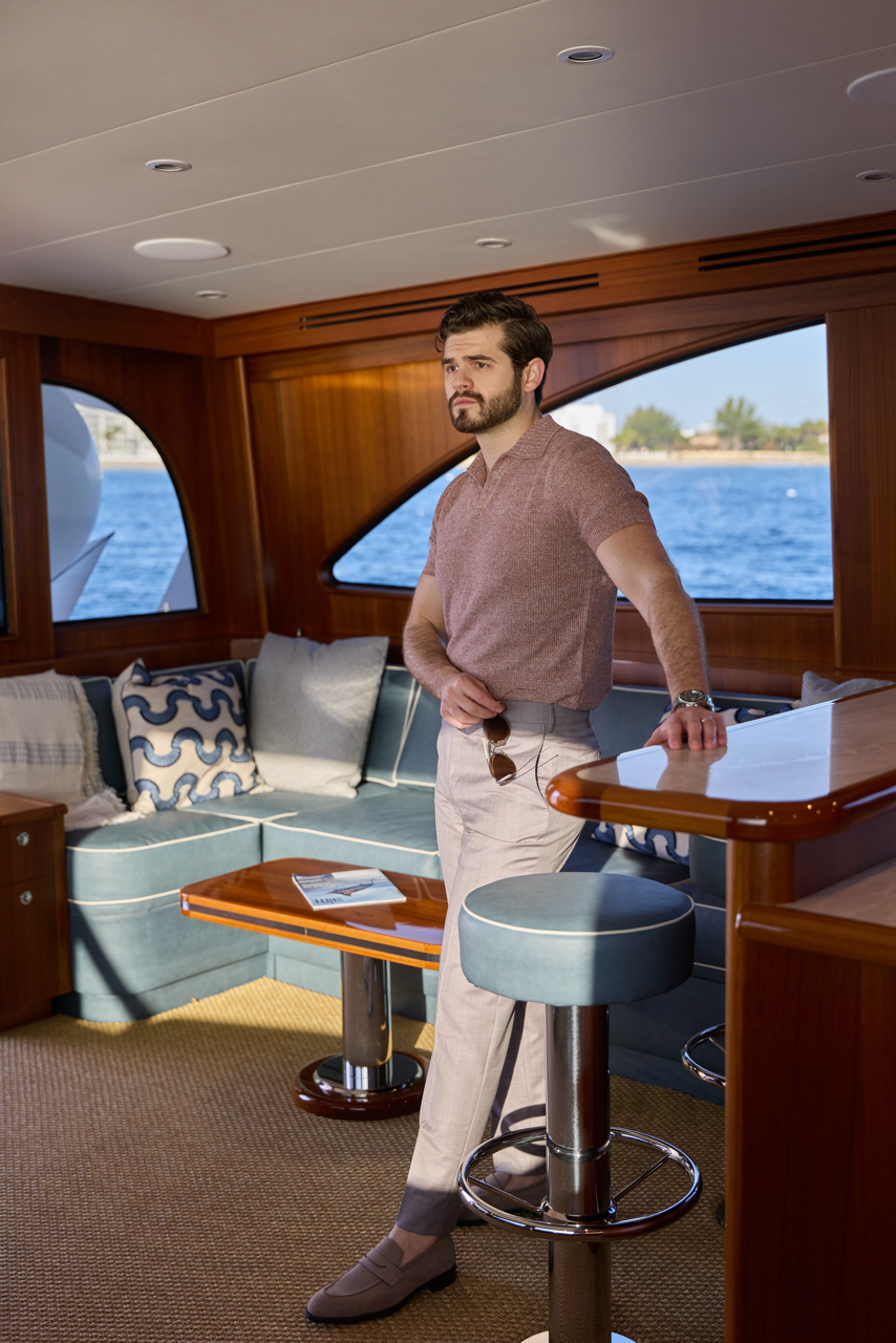 A young man with dark hair and a beard stands in a yacht's interior, looking out the window at the water and shoreline. He is dressed in a short-sleeve brown shirt, light-colored trousers, and gray loafers, holding sunglasses in his right hand with h