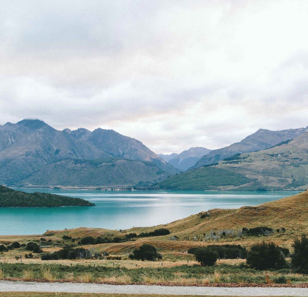Scenic view of a lake with green hills and tall mountains in the background under an overcast sky.