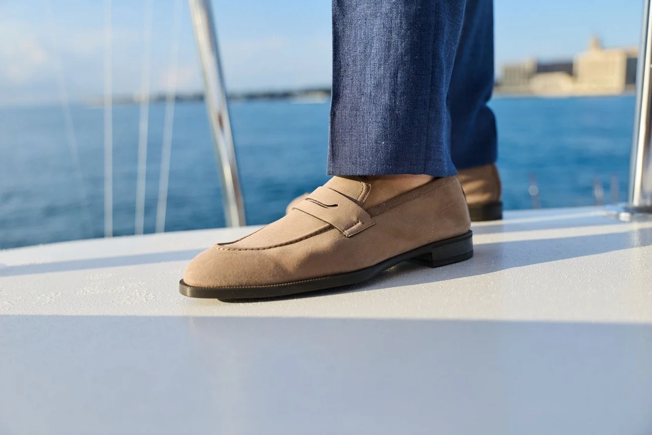 Close-up of a person's feet wearing beige loafers and blue pants, standing on a boat with water and city skyline in the background.