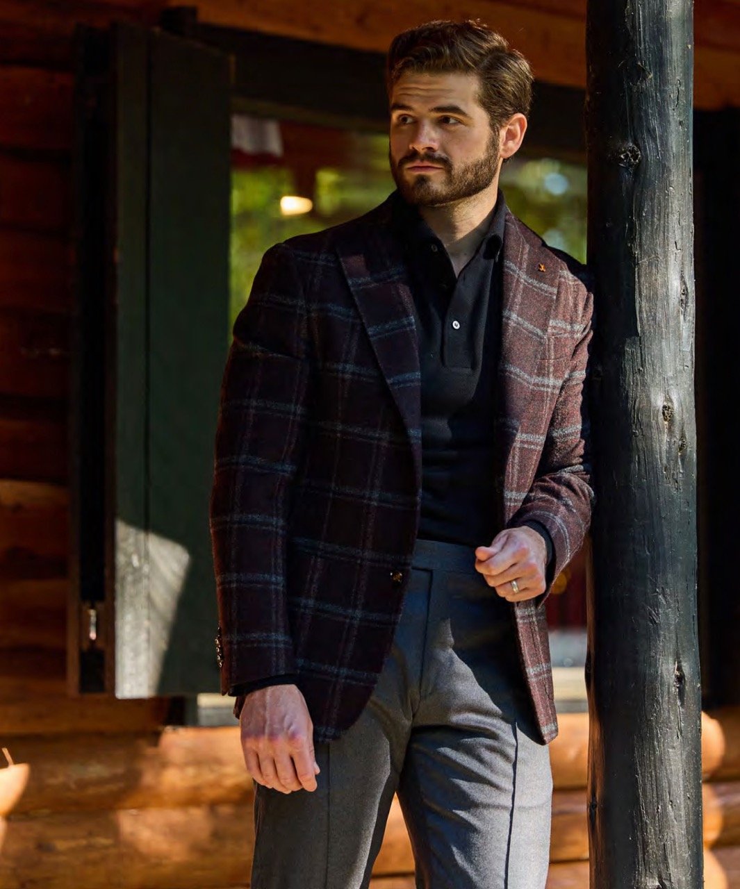A man wearing a dark plaid blazer, black shirt, and gray trousers, standing outdoors near a wooden post.