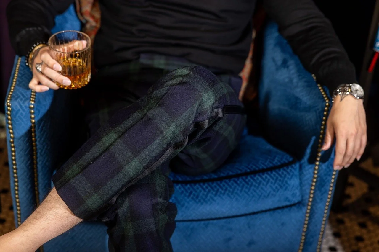 Person sitting on a blue upholstered armchair, holding a single-malt scotch in a rocks glass in their right hand, wearing a traditional Blackwatch plaid in Malcom's Parma Formal trouser model, a black long-sleeve shirt, and a watch and bracelet.