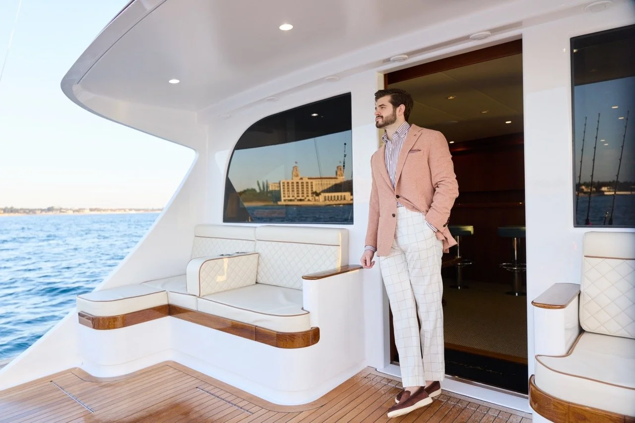 A man in a pink blazer and plaid white pants standing on the deck of a yacht, with water and city buildings in the background.