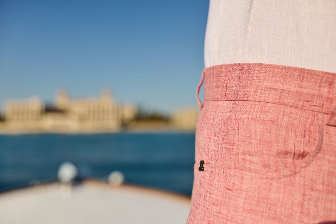 Close-up of a person wearing red shorts and a white shirt, standing on a boat with water and a blurred shoreline in the background.