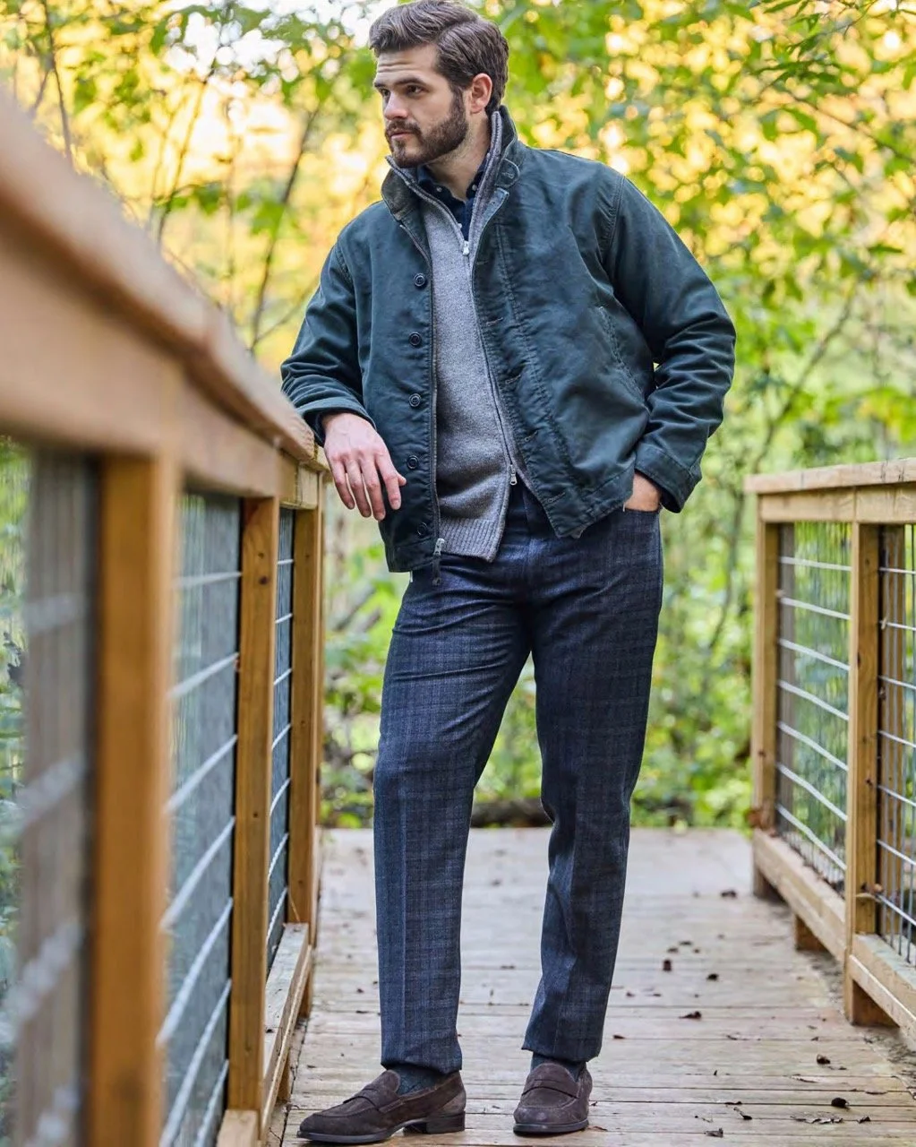A young man with dark hair and beard standing outdoors on a wooden bridge, wearing a dark jacket, gray sweater, dark plaid pants, and brown loafers, with trees and foliage in the background.