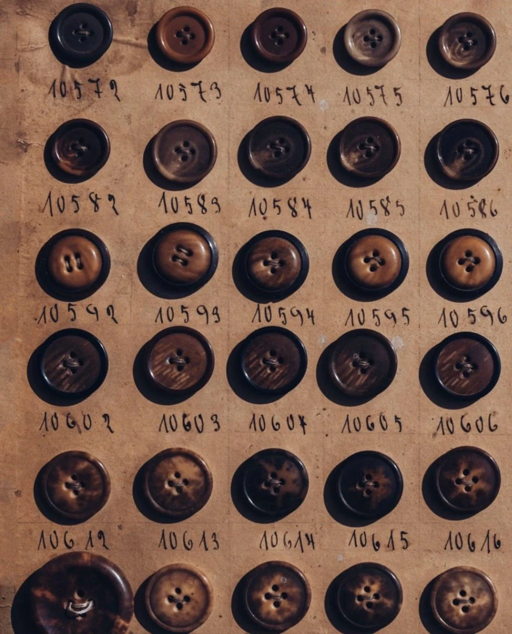 A display of vintage horn buttons arranged in a grid on a cardboard background. Each button has handwritten labels with numbers below them.