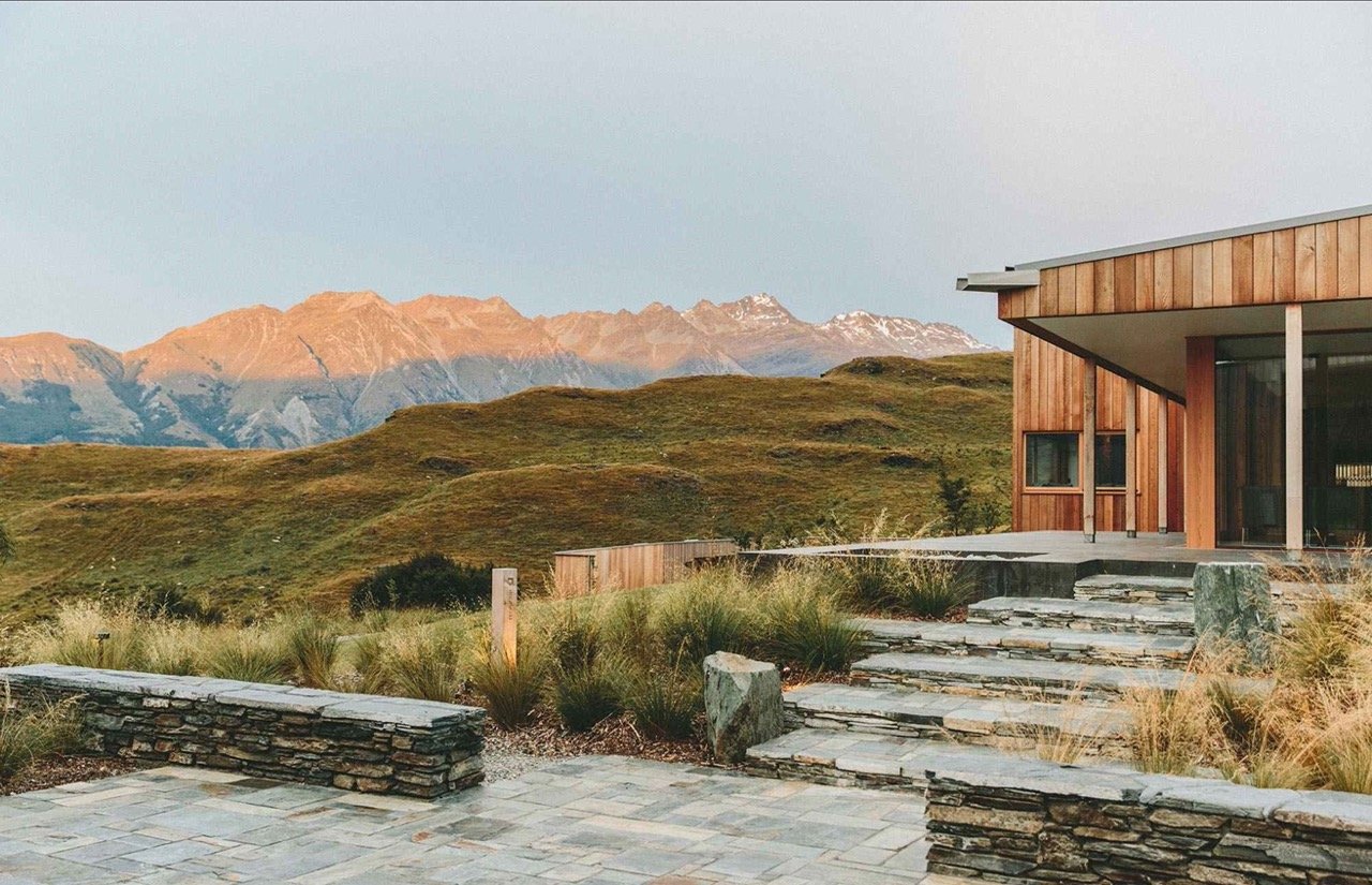 A modern wooden house with stone steps leading to it, surrounded by grassy landscaping and mountains in the background.