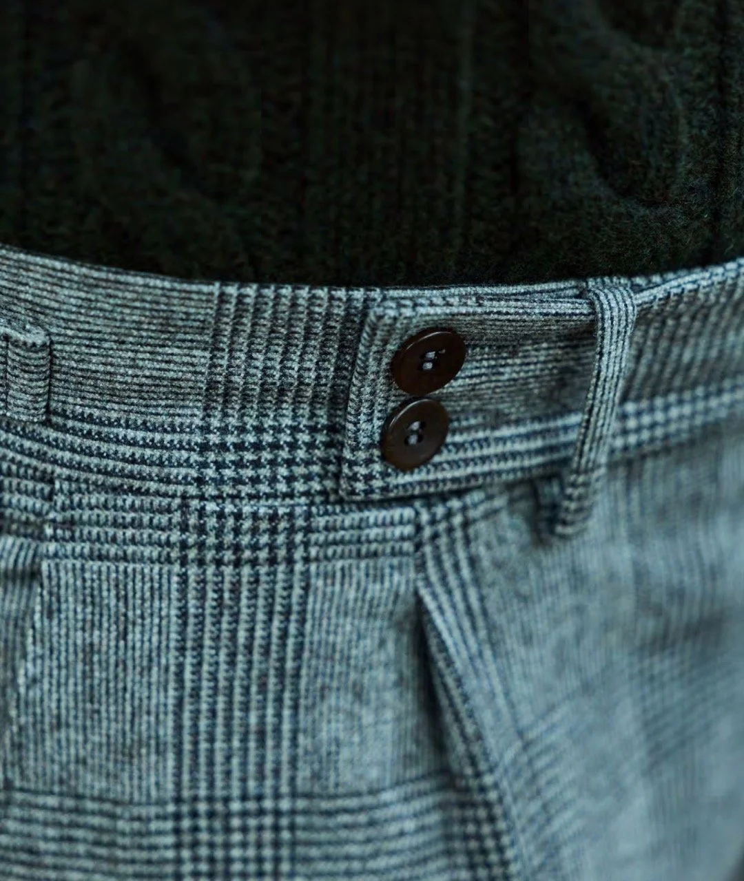 Close-up of a pair of gray houndstooth patterned pants with a waistband featuring two black buttons, worn over a black knit sweater.