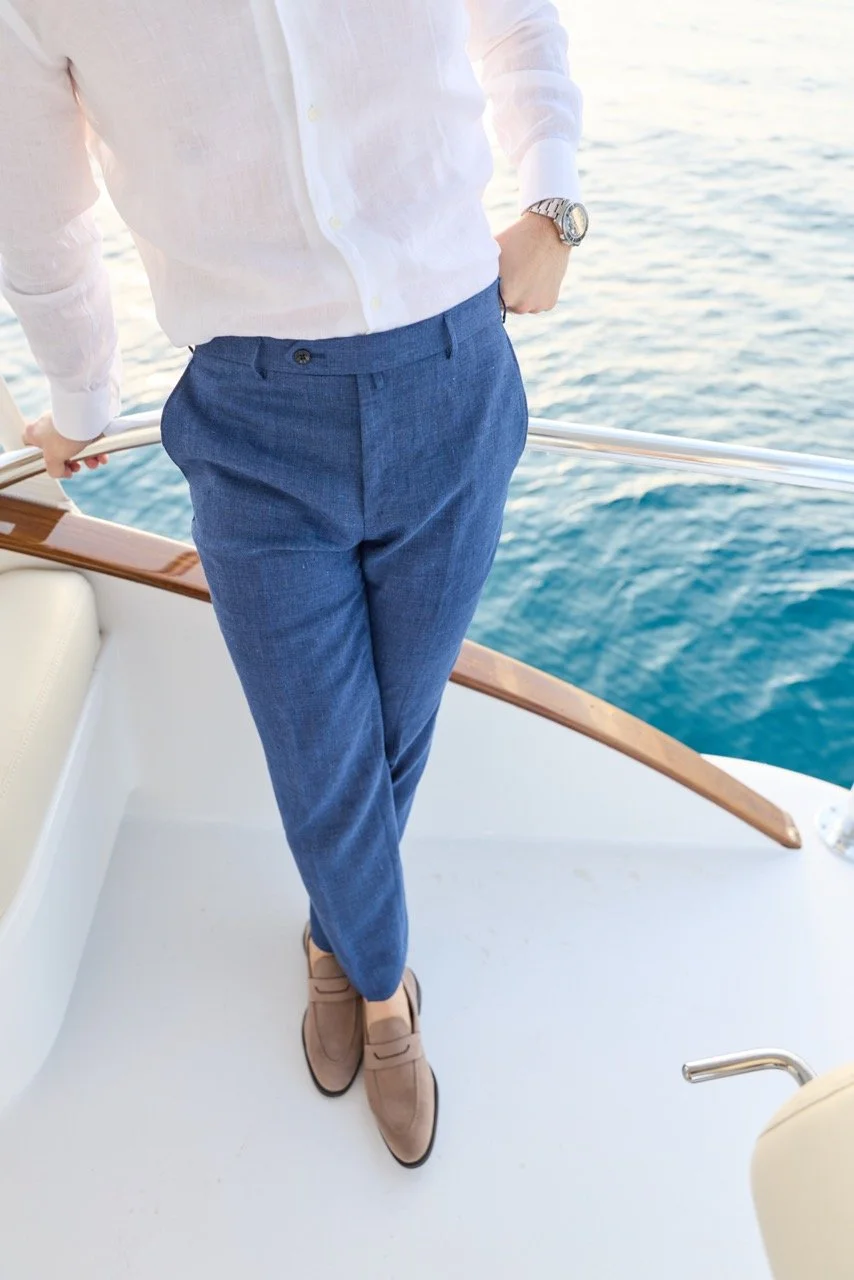 A man standing on a boat railing overlooking the water, dressed in a white shirt, blue trousers, tan shoes, wearing a wristwatch.