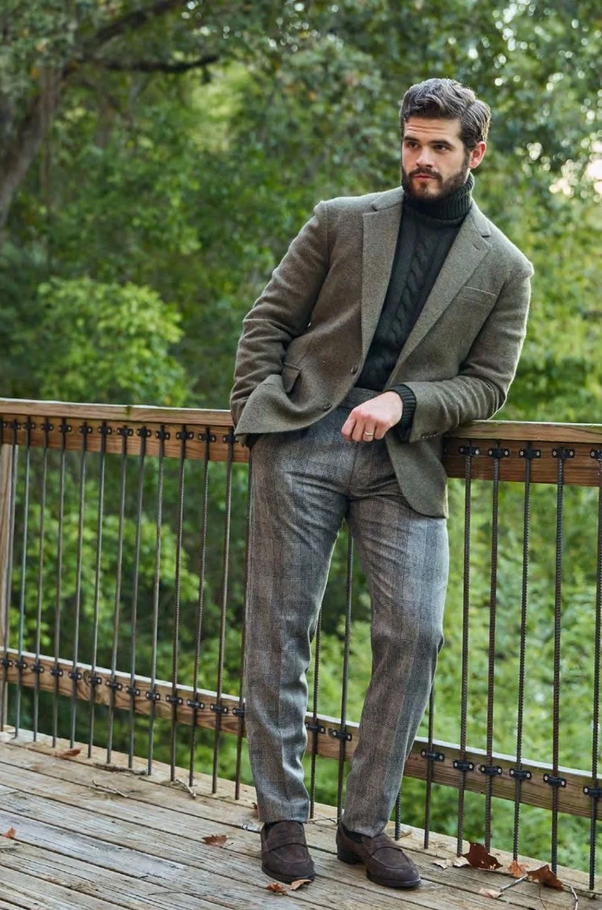 A man with dark hair and a beard standing on a wooden balcony outdoors, wearing a brown blazer, black turtleneck sweater, gray plaid pants, and dark shoes, with lush green trees in the background.
