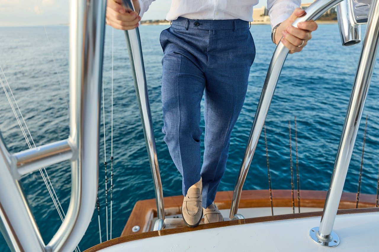 A person in formal attire, including beige shoes and blue pants, is standing on the deck of a boat, holding onto the metal railing, with a view of the water and the shoreline in the background.