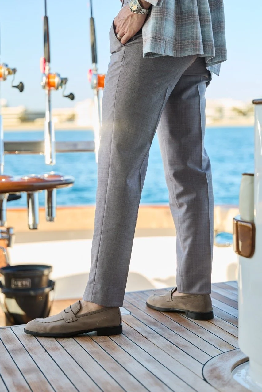 Close-up of a man in gray dress pants, beige shoes, and a plaid blazer standing on a boat deck by the water, with fishing rods in the background.