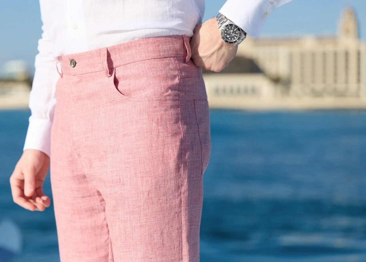 Close-up of a man's pink linen pants and part of his white shirt, with a wristwatch on his wrist, standing outdoors near water, with buildings in the background.