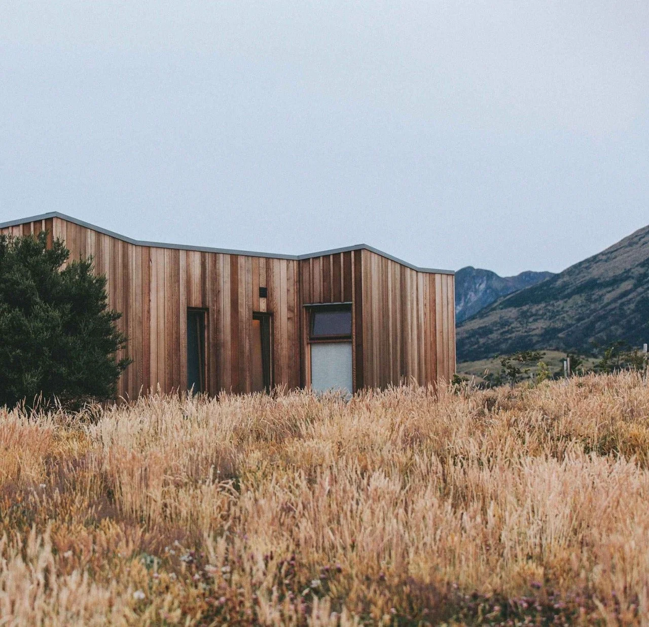 A modern wooden house with a unique roof design situated in a grassy field with mountains in the background under a clear sky.