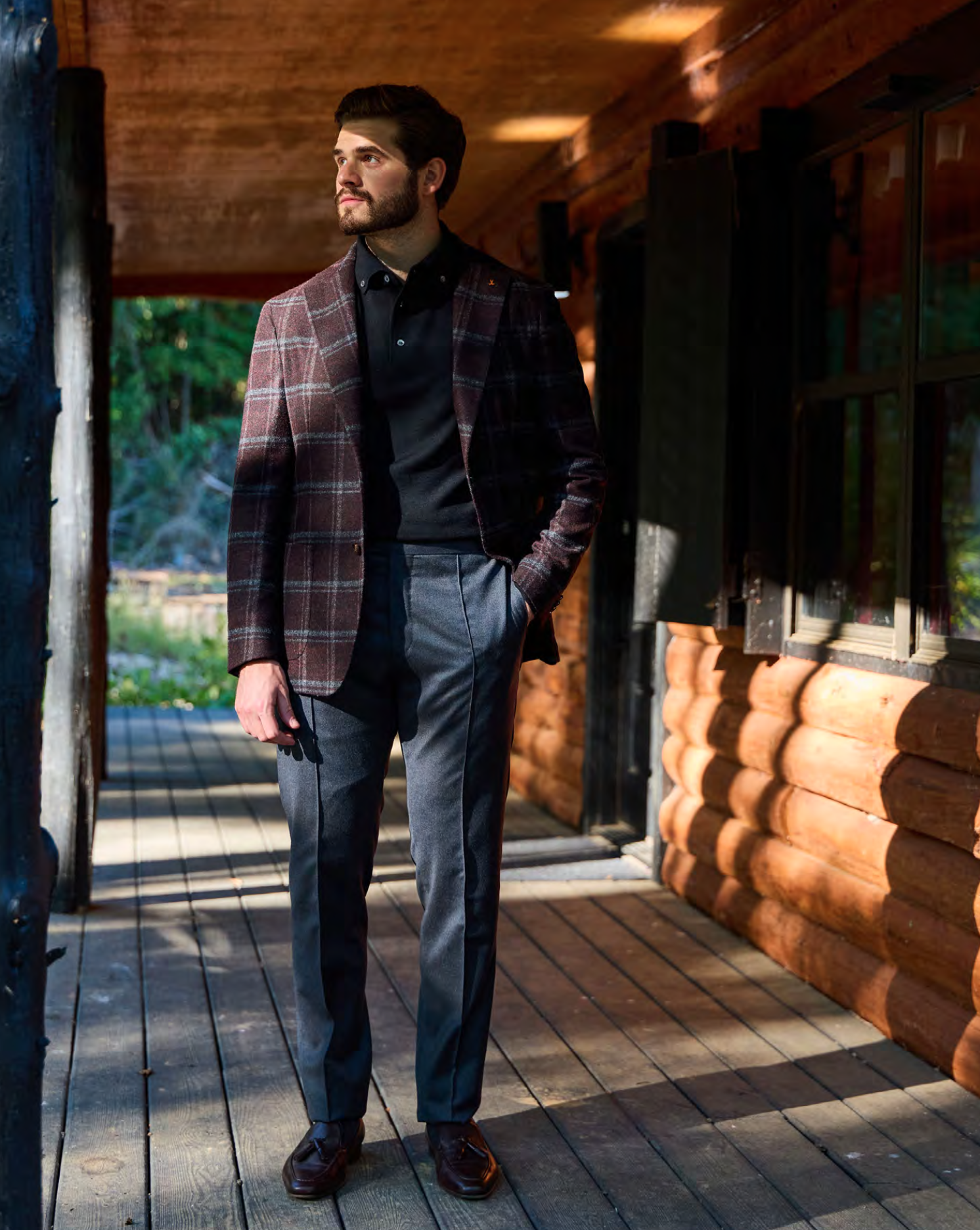 A man dressed in Malcom's Biella model trousers,  a black shirt and plaid blazer standing on a wooden porch, with a log cabin in the background, looking to his right..