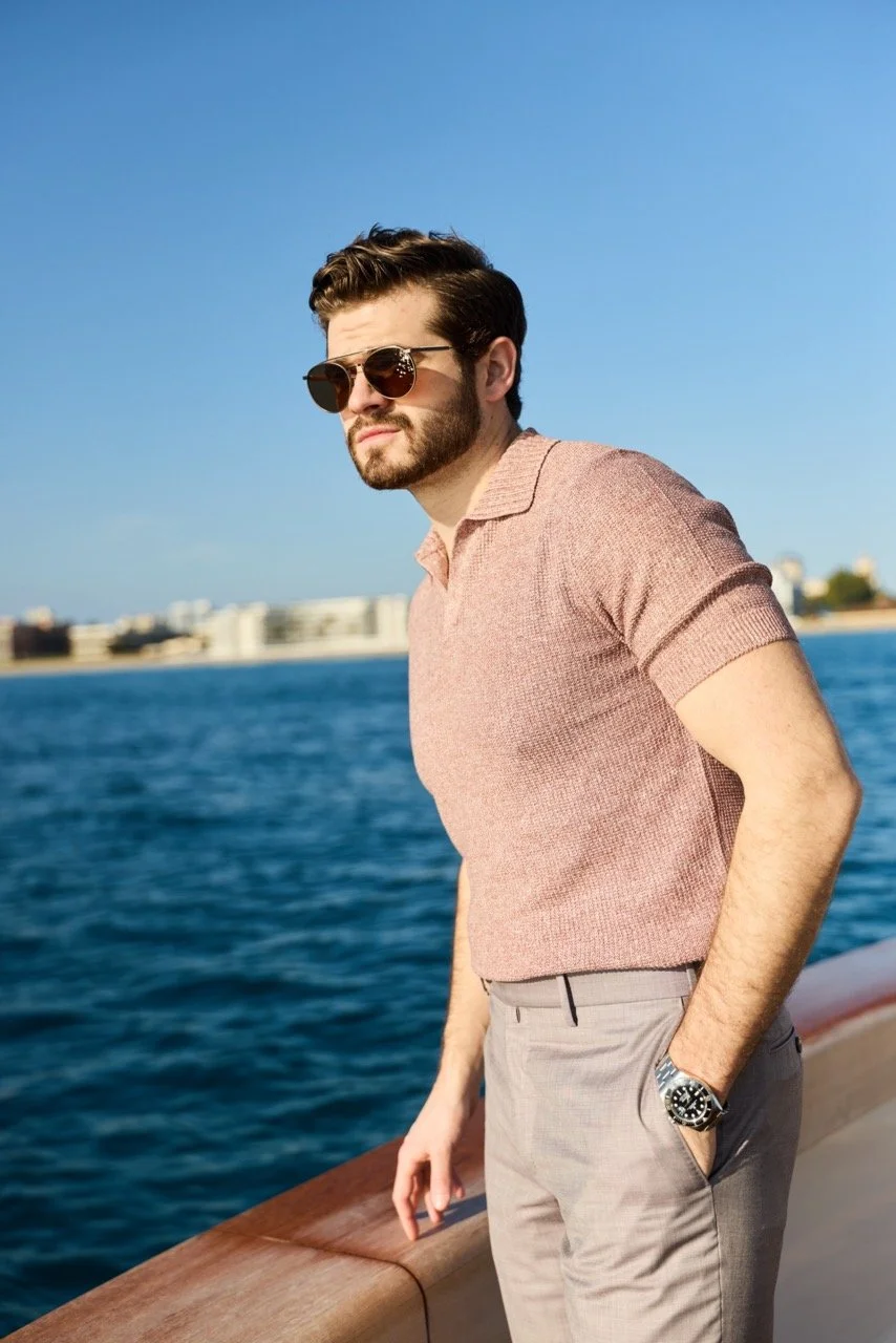 Man with brown hair, beard, sunglasses, wearing a pink polo shirt and gray pants, standing on a boat near water with buildings in the background.