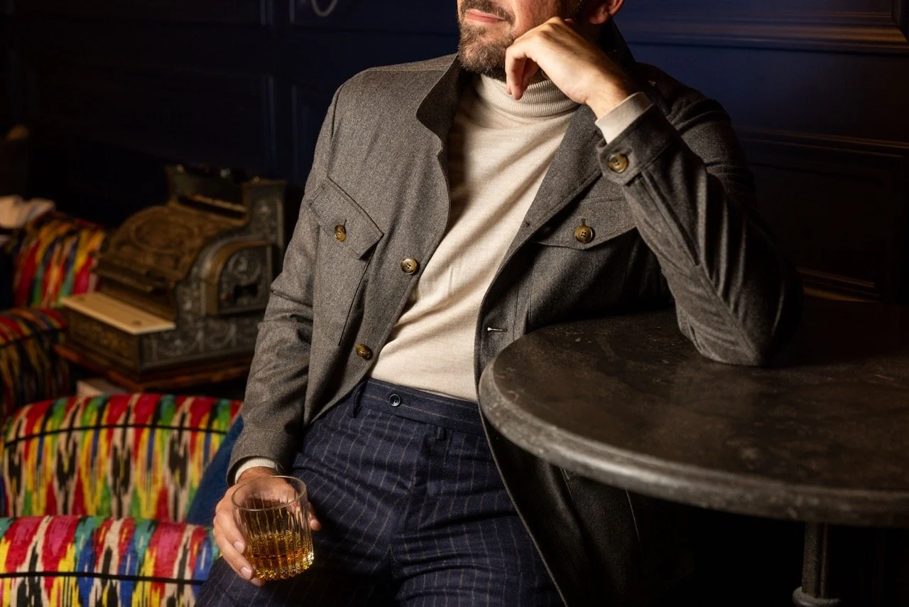 Man with a beard in a gray jacket and turtleneck holding a glass of whiskey, leaning on a dark round table in a dimly lit room with colorful patterned chairs and vintage decor in the background.